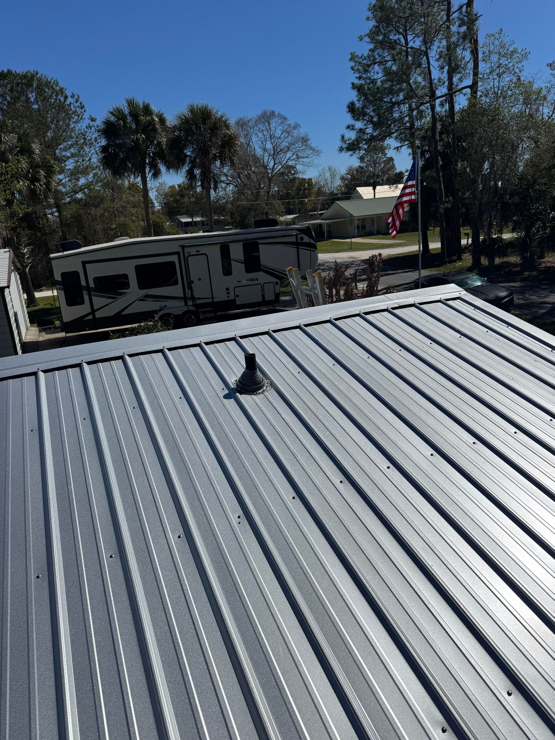 A metal roof with a plumbing vent boot, with trees, a camper, and an American flag visible in the background.