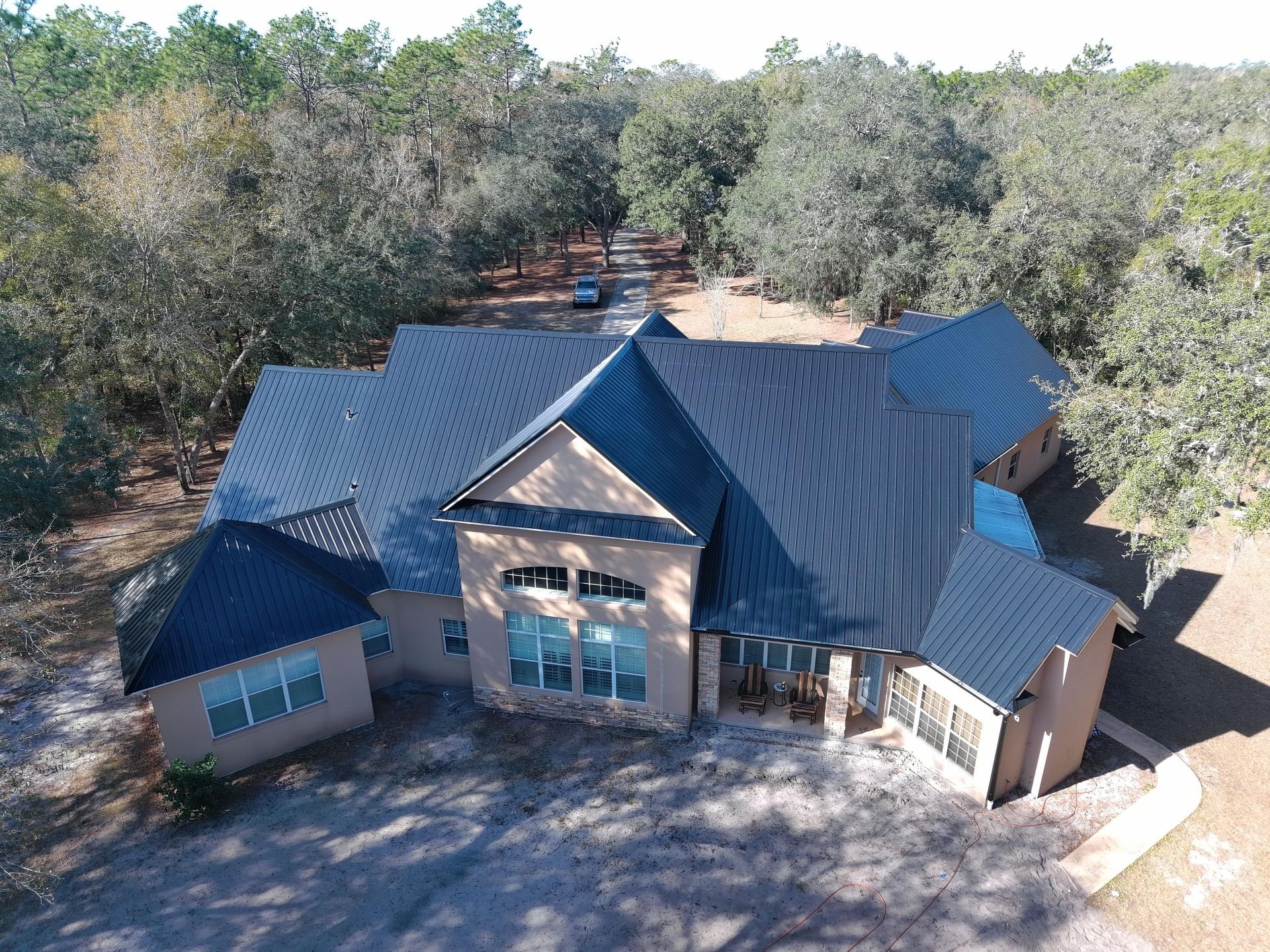 Aerial view of a tan, single-story house with a dark metal roof surrounded by woods and a paved driveway.