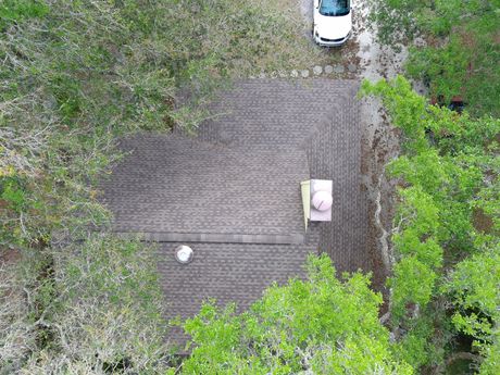 A brown single-story house with a gray roof and a tan garage door sits behind a gravel driveway under a clear blue sky.