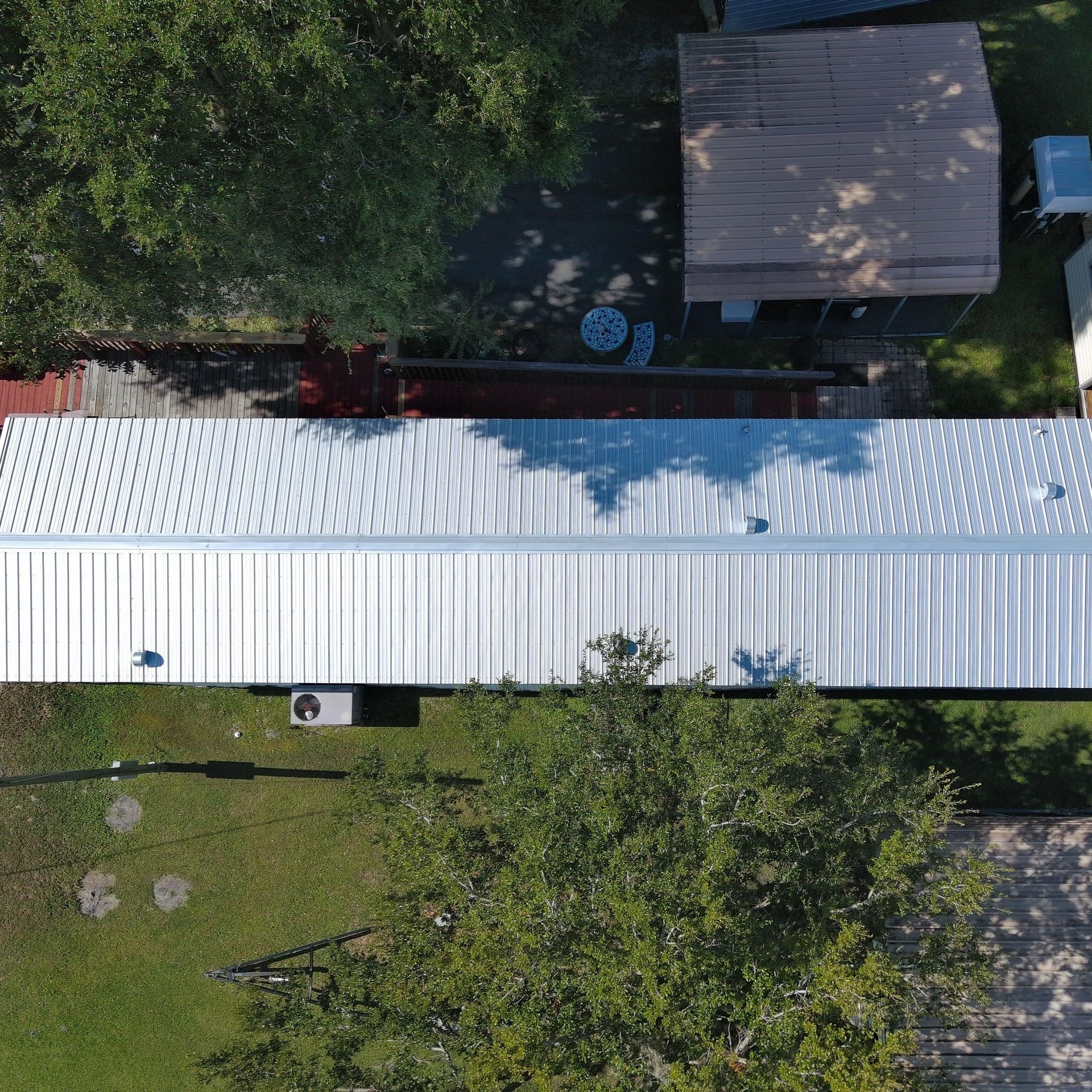 An aerial view of a long, rectangular home with a silver metal roof, surrounded by green grass and trees.