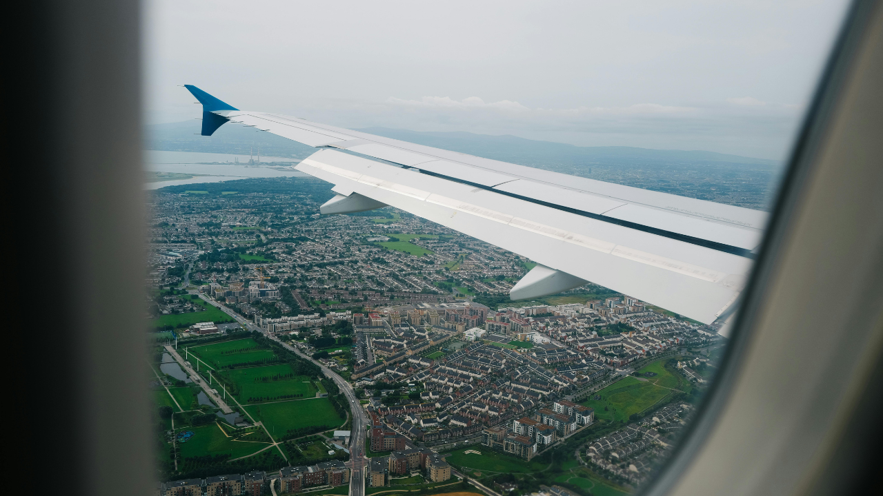 View from an airplane window, showing a wing and cityscape with green spaces. Cloudy sky.