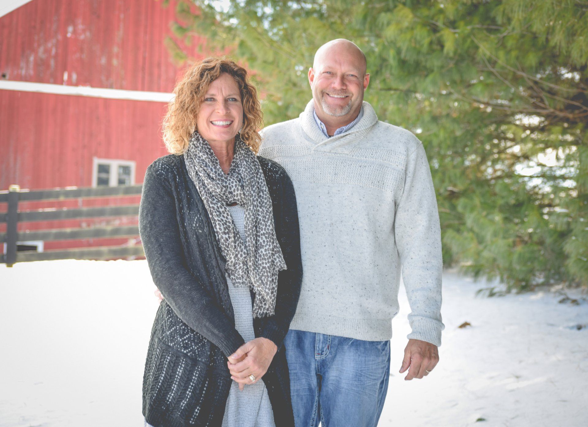 Couple standing in snow in front of a red barn, woman wearing scarf and cardigan, man in sweater.