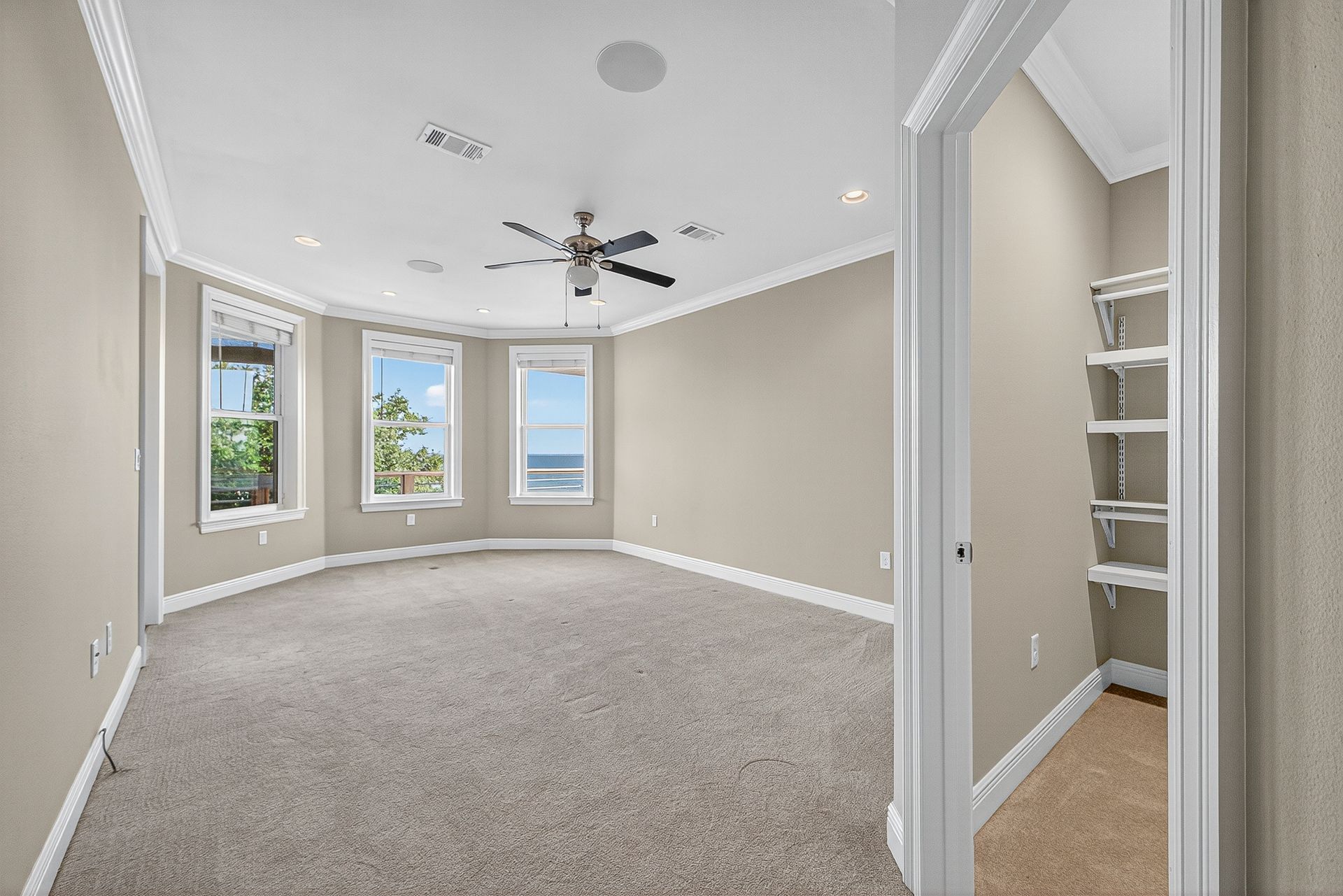 Empty beige bedroom with carpet, ceiling fan, three windows, and a built-in shelf nook