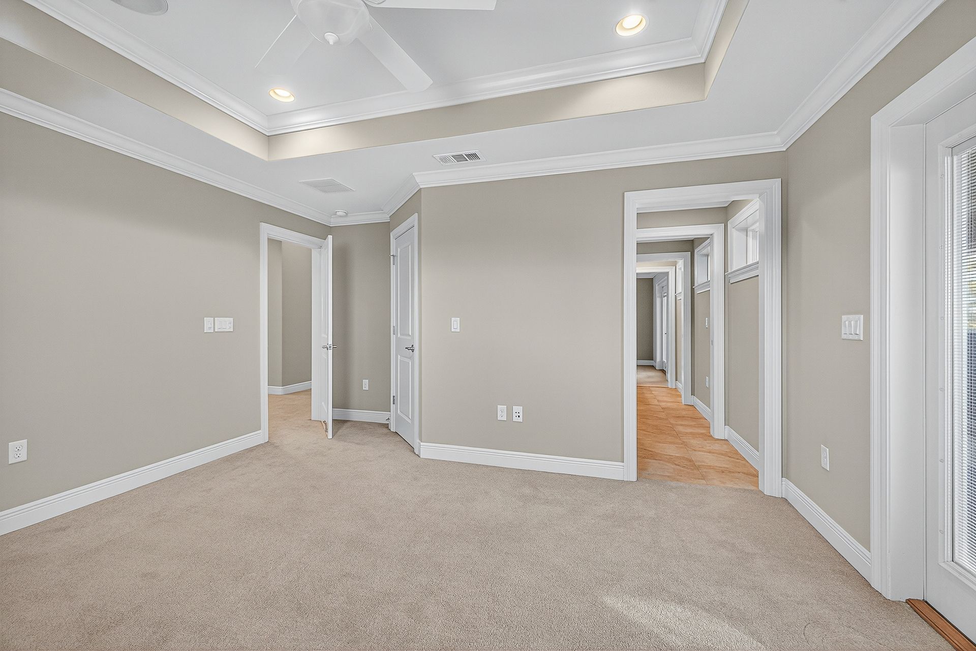 Spacious beige carpeted room with gray walls, tray ceiling, and open doorways to adjacent rooms.