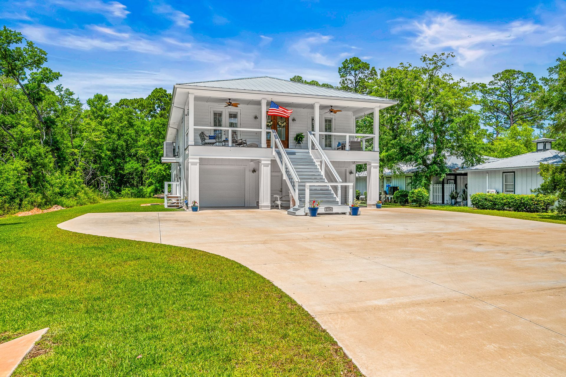 Modern white two-story house with exterior stairs and a large concrete driveway surrounded by green trees.