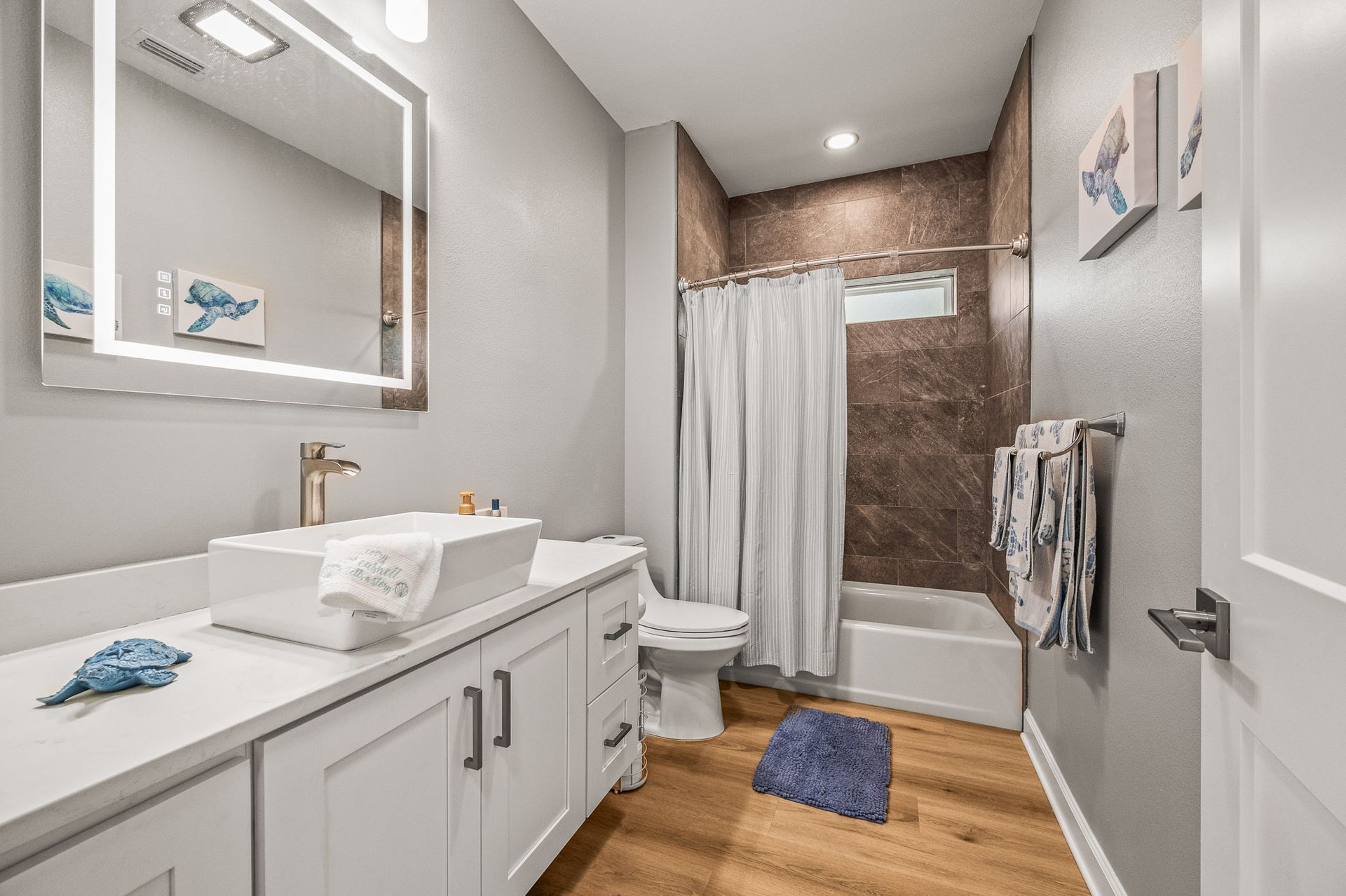 Bright modern bathroom with white vanity, mirror, shower curtain, toilet, and blue rug on wood floor.