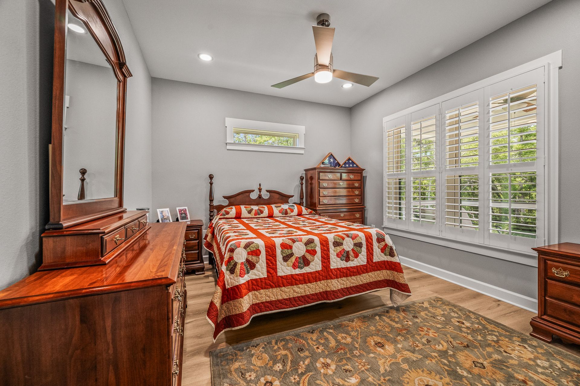 Bedroom with red patchwork quilt, wooden dresser, large mirror, ceiling fan, and bright plantation shutters
