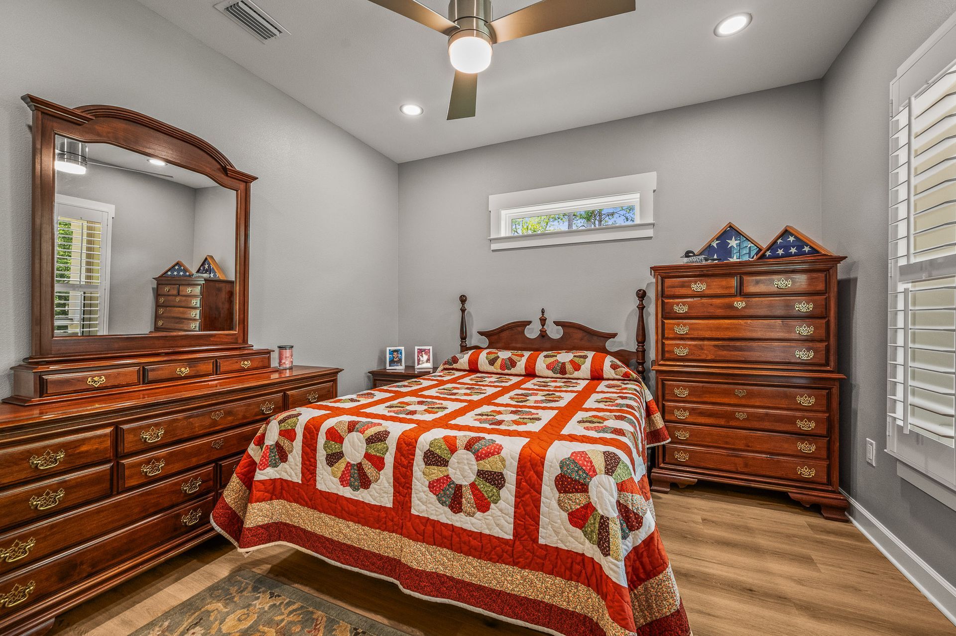 Bedroom with red-and-green quilt, wooden dresser and mirror, and a bed beneath a ceiling fan.
