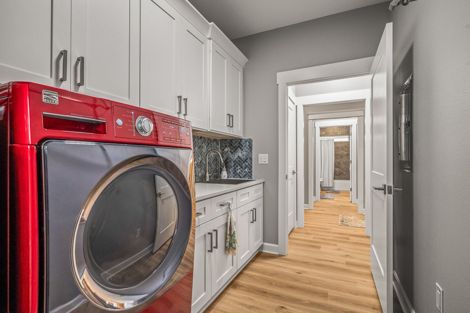 Bright laundry room hallway with red washer, white cabinets, gray walls, and wood floors