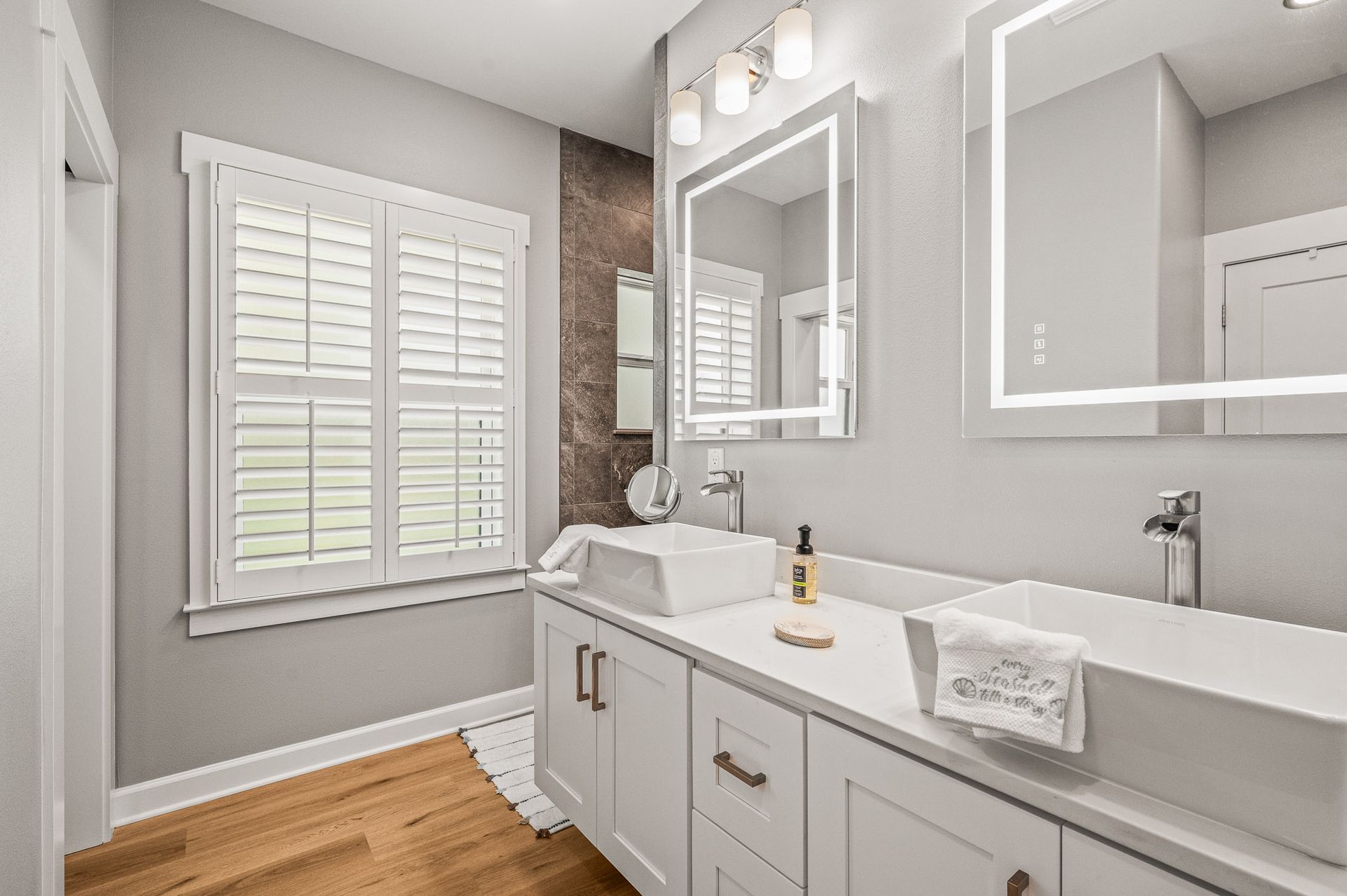 Bright modern bathroom with white vanity, mirror, sink, and window shutters over wood floor.