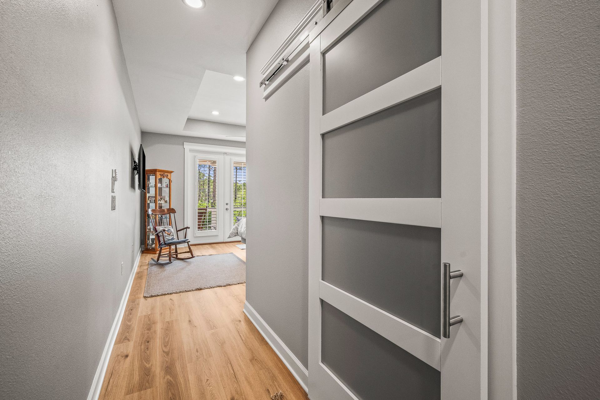Modern hallway with wood floors, white walls, and a gray door leading to a bright room with a ladder inside.