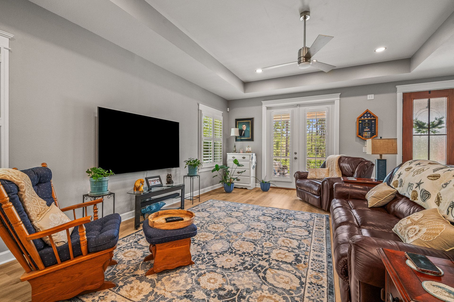 Bright living room with leather and wooden chairs, patterned rug, wall-mounted TV, and large windows.