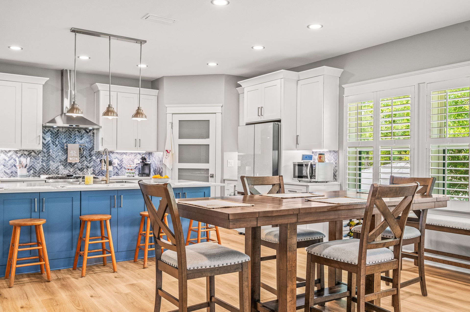 Bright kitchen and dining area with blue island, white cabinets, wooden table, stools, and large windows