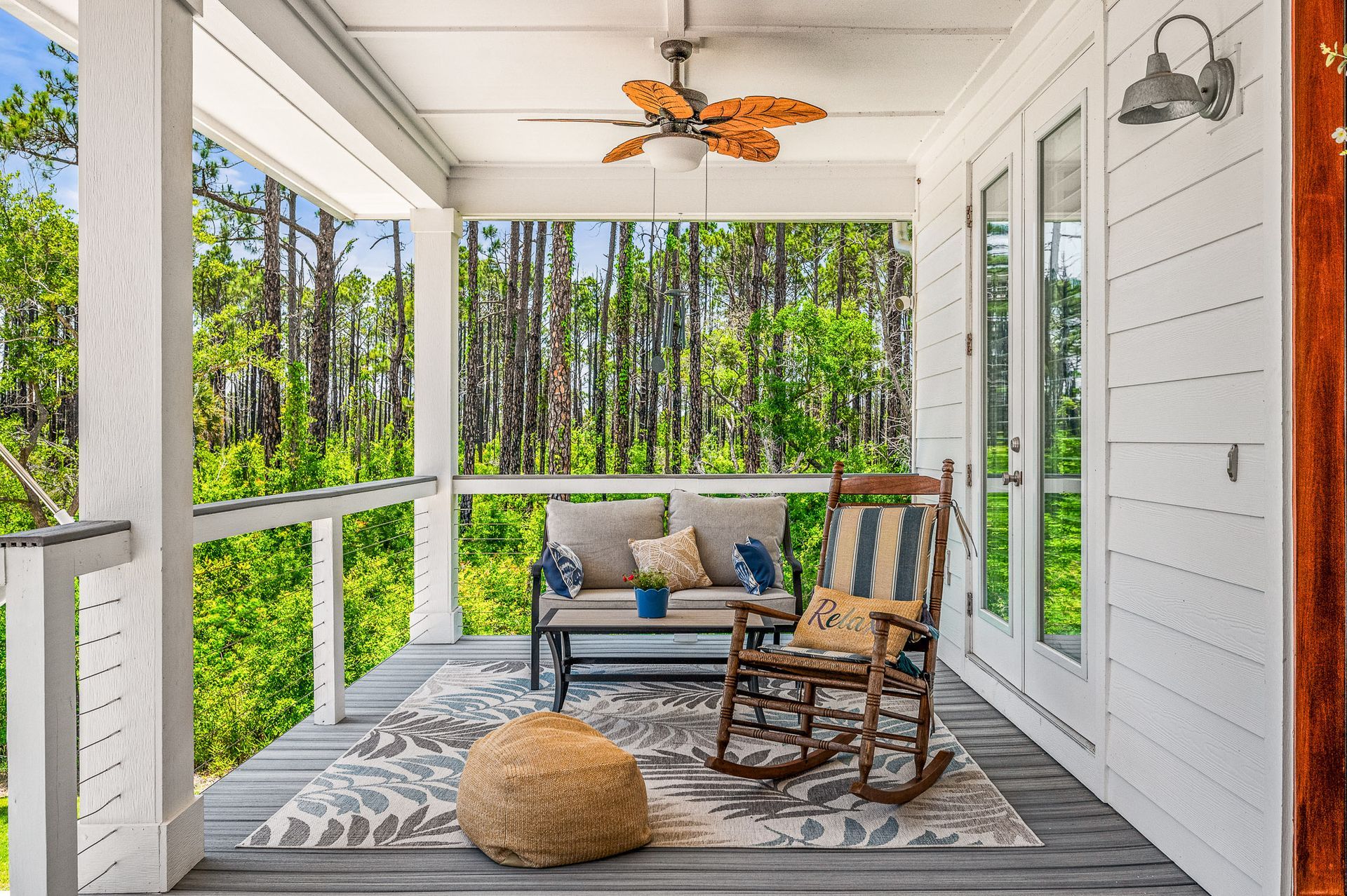 Screened porch with gray rug, blue bench, rocking chair, and wicker ceiling fan overlooking green woods