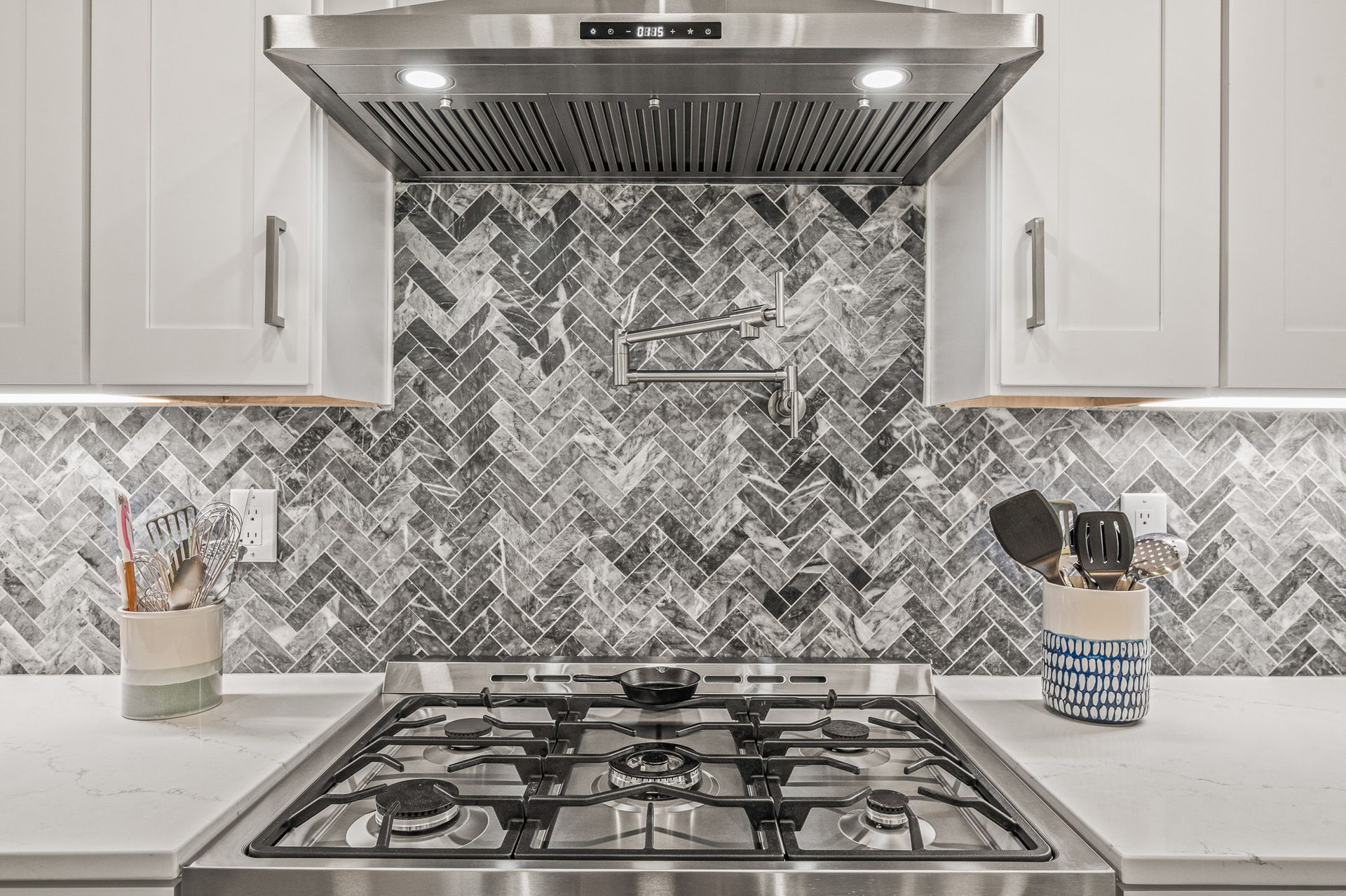 Modern kitchen stove with stainless hood, white cabinets, and gray herringbone tile backsplash