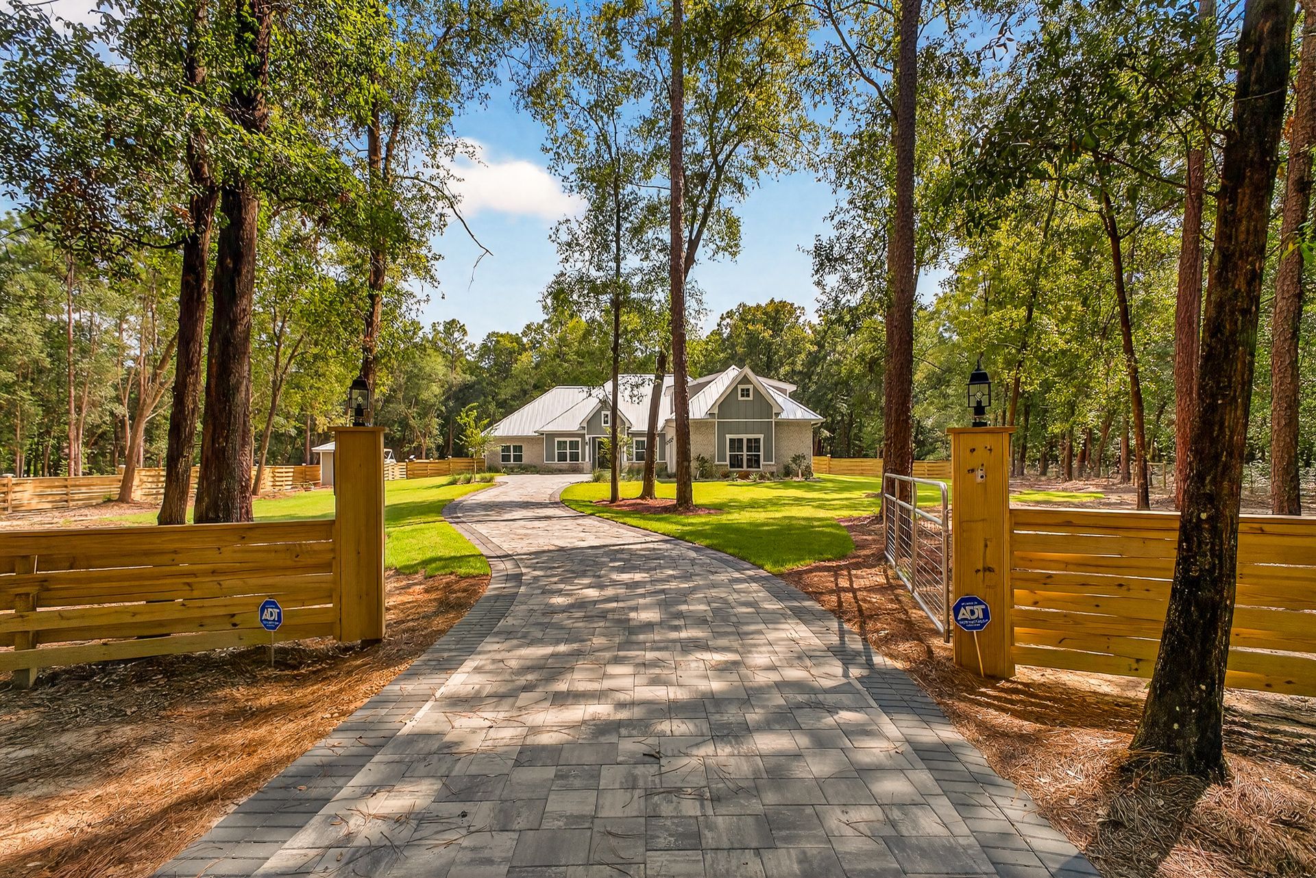 Paved driveway through tall trees leading to a white house with wooden fence posts on both sides