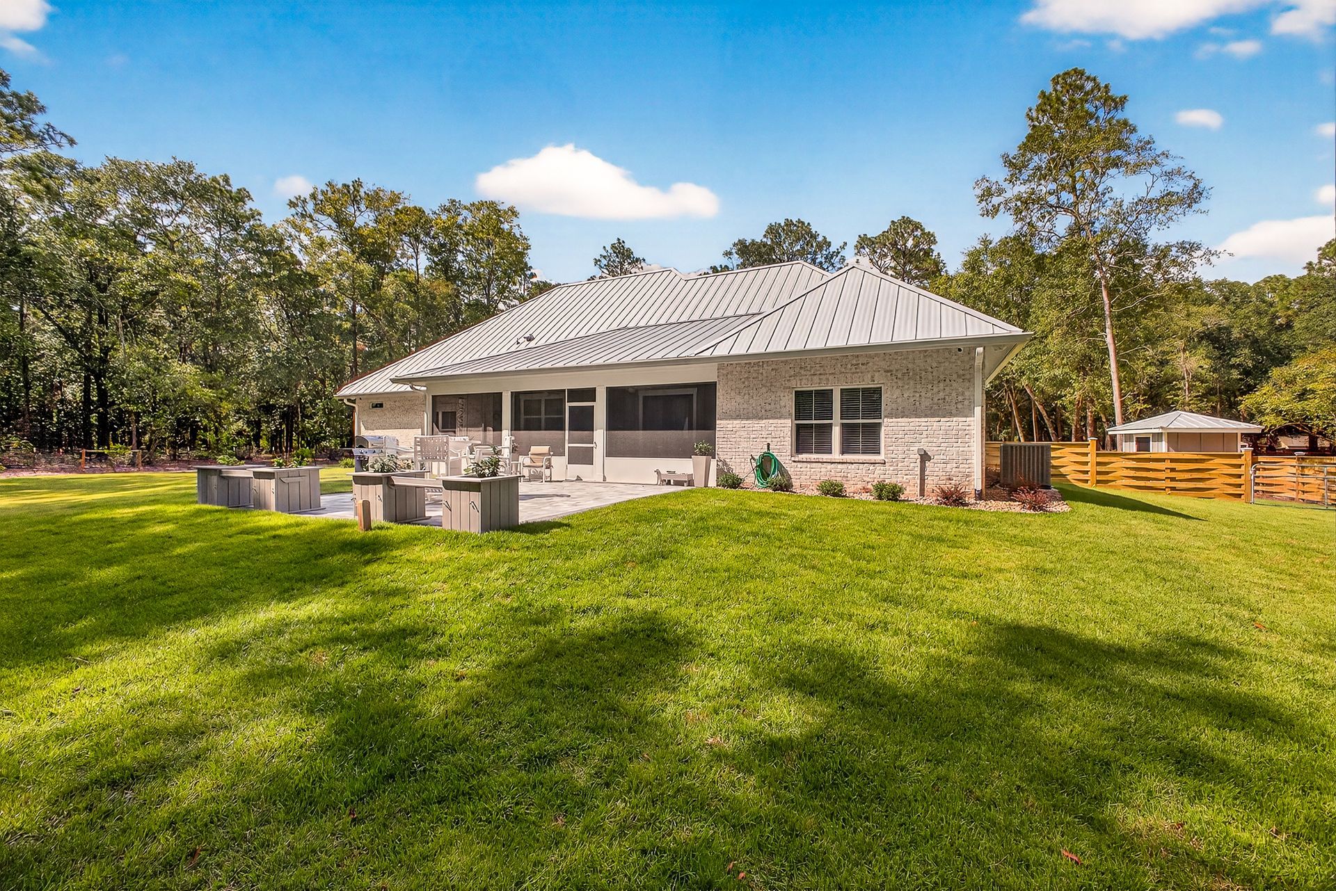 Stone house with a gray roof and patio, set in a sunny grassy yard with trees and blue sky.