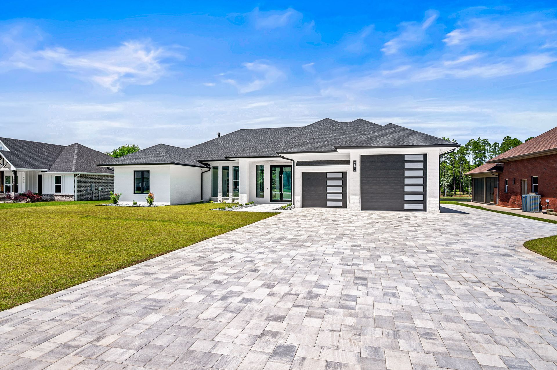 Modern single-story house with a gray roof, large paver driveway, and green lawn under a blue sky