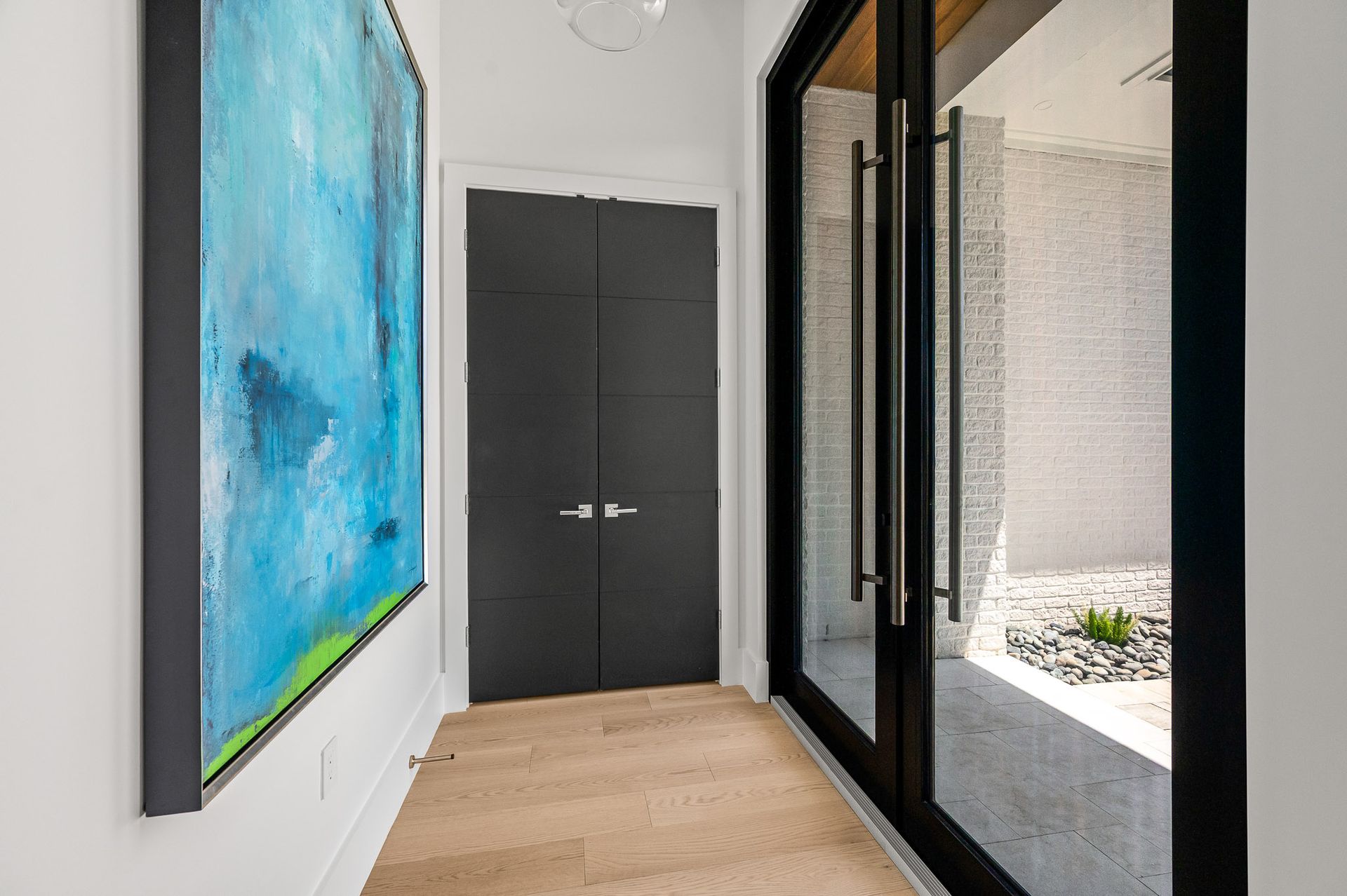 Modern hallway with blue abstract art, dark double doors, and glass wall overlooking a small courtyard.