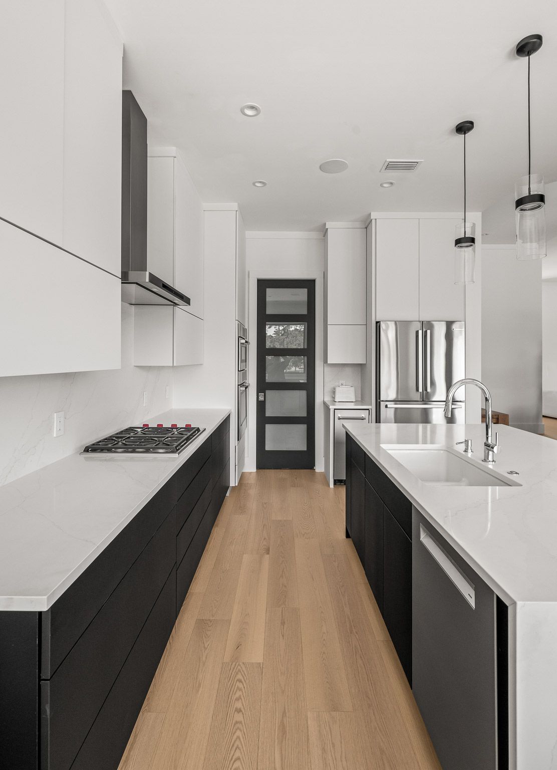 Modern white kitchen with black cabinets, wood floor, island sink, and a door at the end.