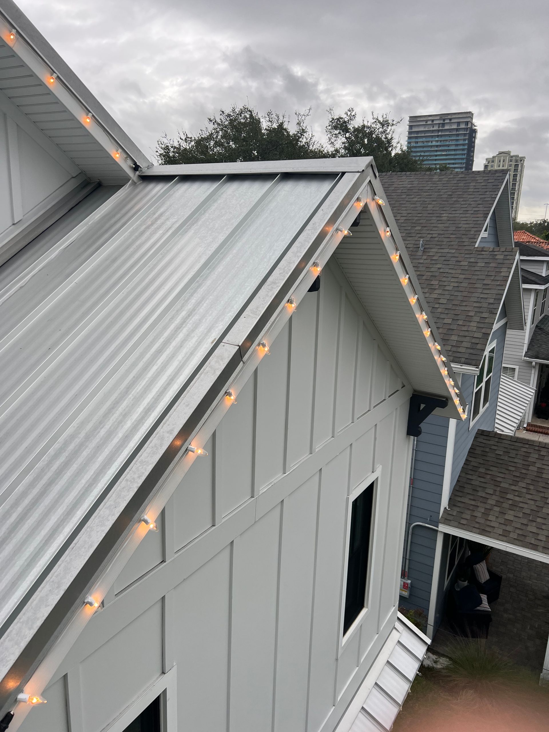 Gray house with metal roof, string lights, and vertical siding. Other similar houses in background.