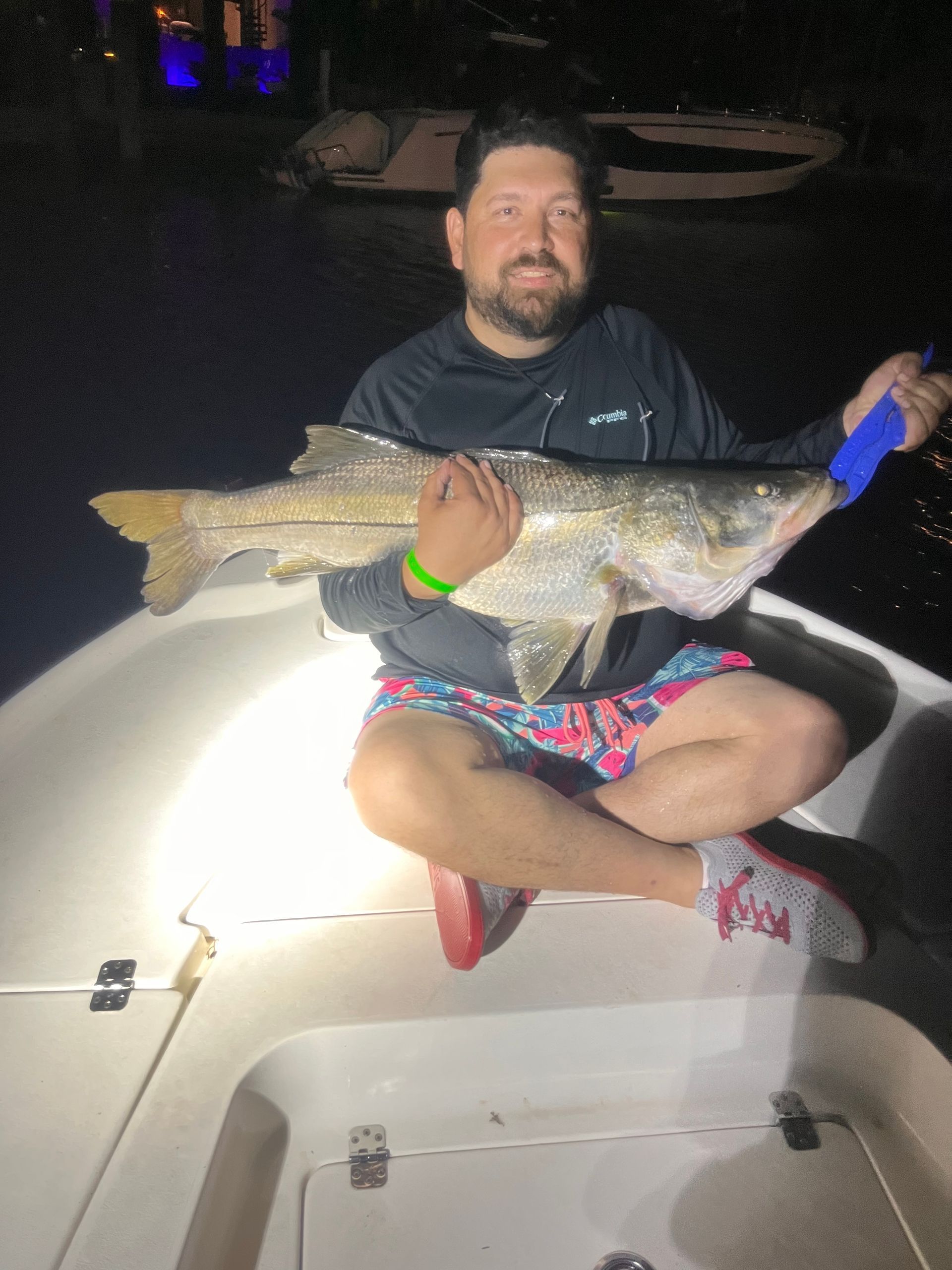 Man sitting on a boat at night holding a large fish, illuminated by a bright light