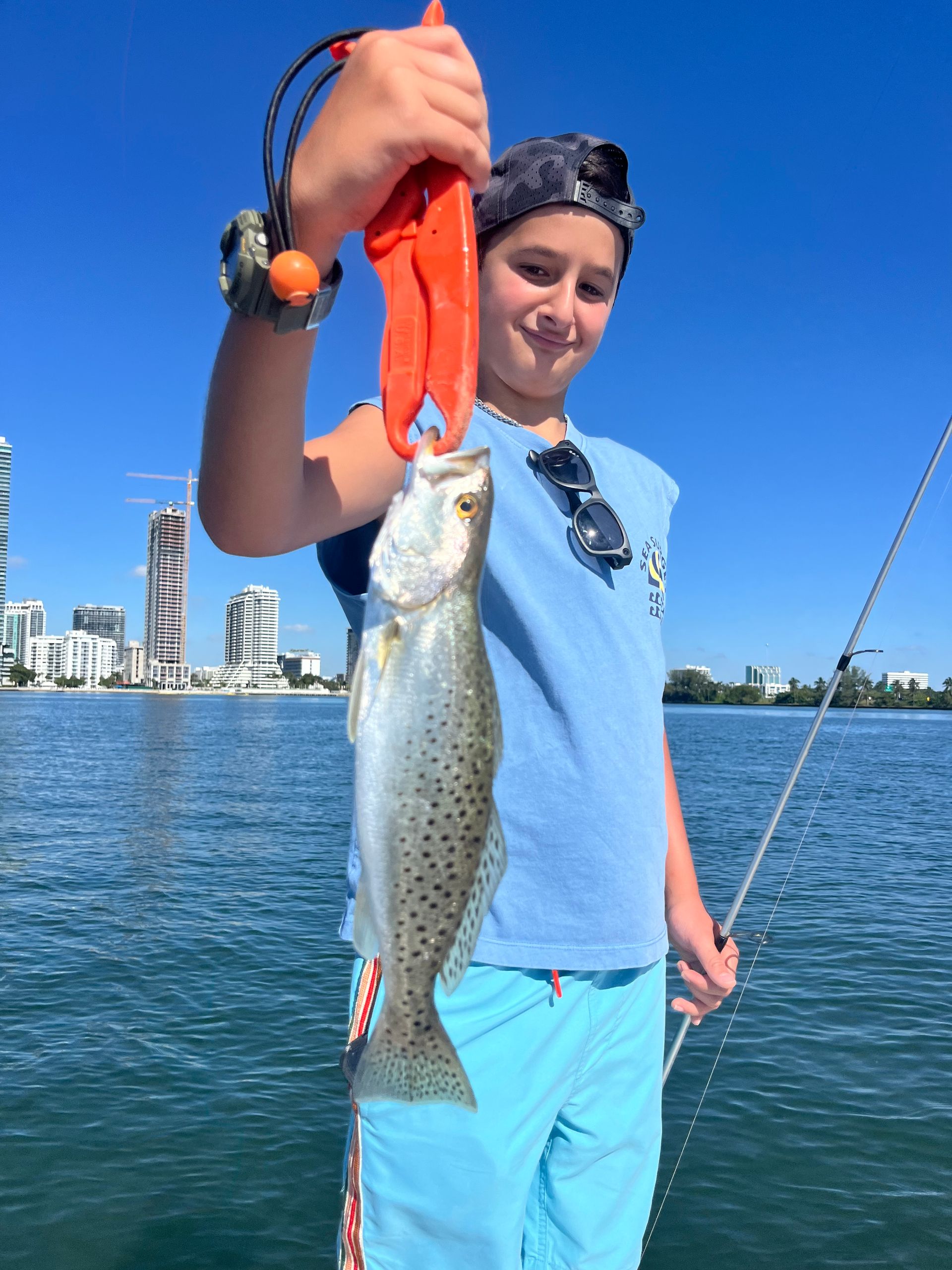 Person holding up a fish on a bright blue waterfront, wearing a light blue shirt and shorts.
