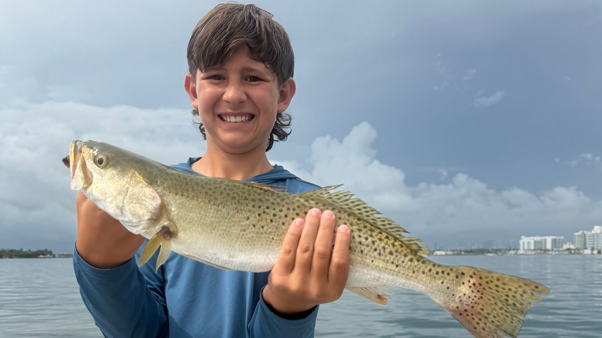 Smiling person holding a large speckled fish by the water with cloudy sky and distant buildings