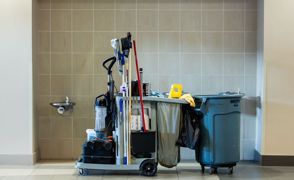 Cleaning cart with mops and supplies beside blue trash bins in a tiled utility room
