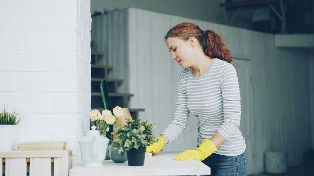 Woman in yellow gloves arranging flowers at a kitchen counter beside a white brick wall.