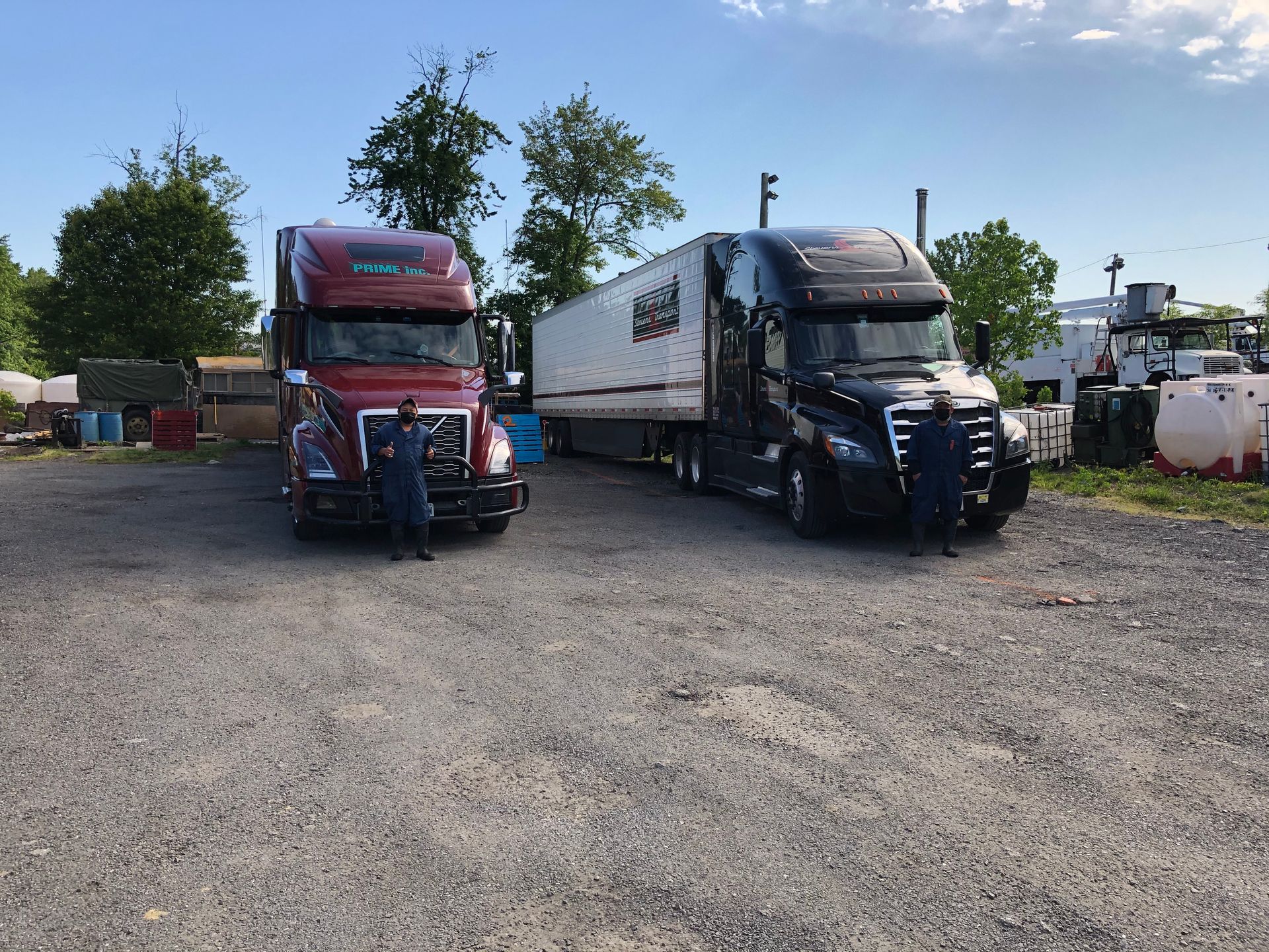Two semi-trucks parked on gravel. One is red, the other black with a trailer. Two people stand near the trucks.