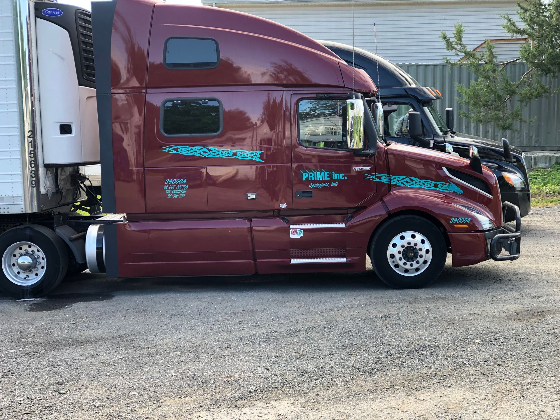 Two parked semi-trucks, one maroon with teal lettering, the other black, on a gravel lot.