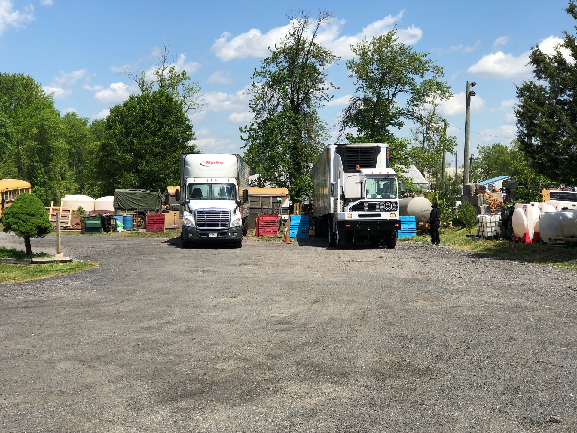Two trucks parked on gravel, with trees and various containers in the background under a blue sky.