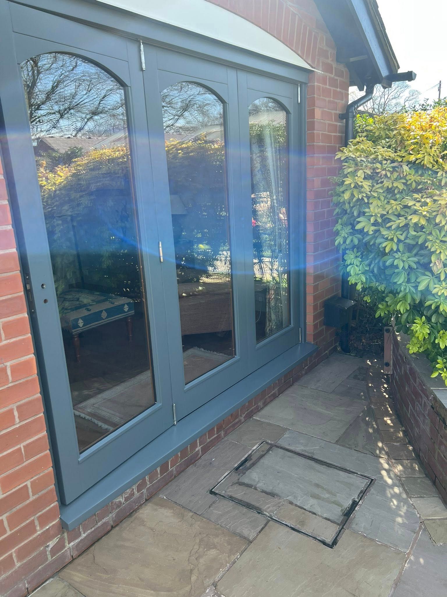 Brick patio with gray French doors, reflecting trees and sky, beside a bush.
