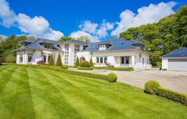 Large beige two-story house with dark roof, front porch, and landscaped driveway surrounded by trees