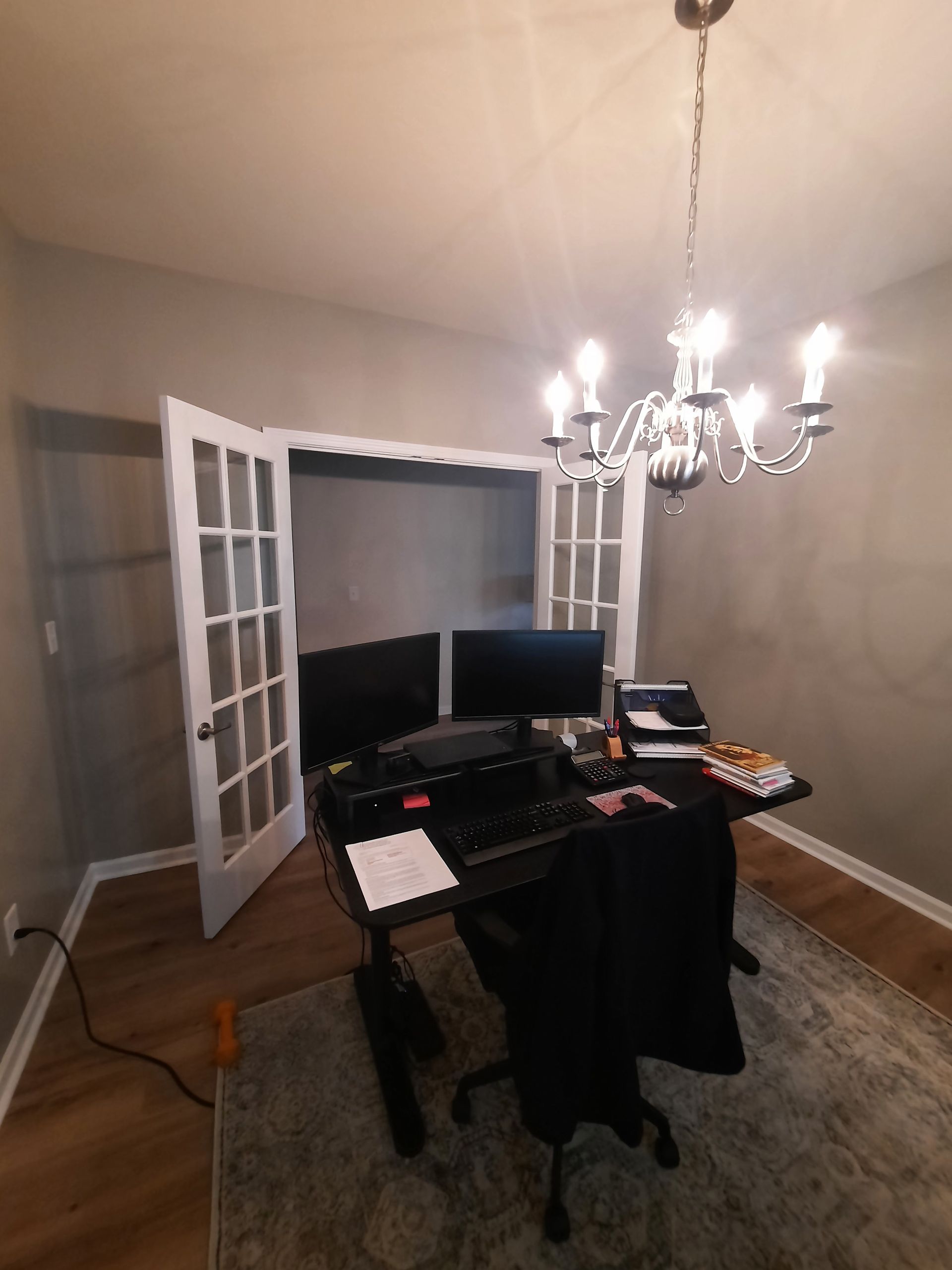 A room with a desk and two computer monitors and a chandelier hanging from the ceiling.