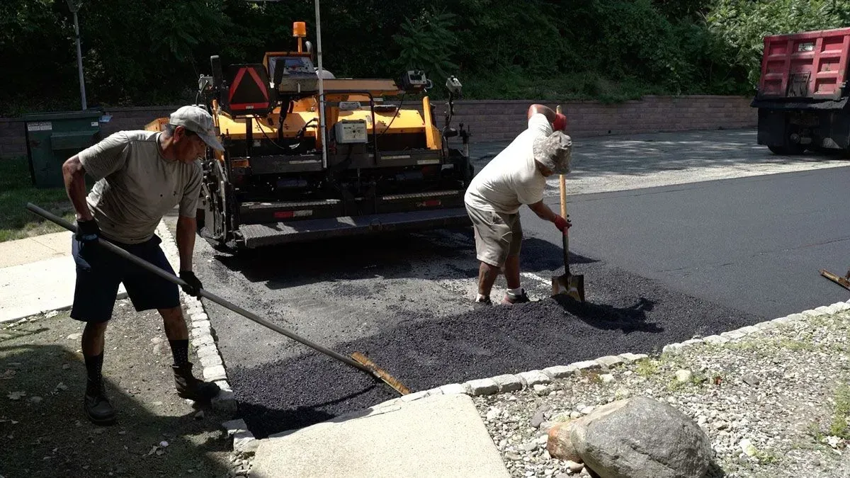 Two road workers paving a street with asphalt; one rakes, the other shovels.