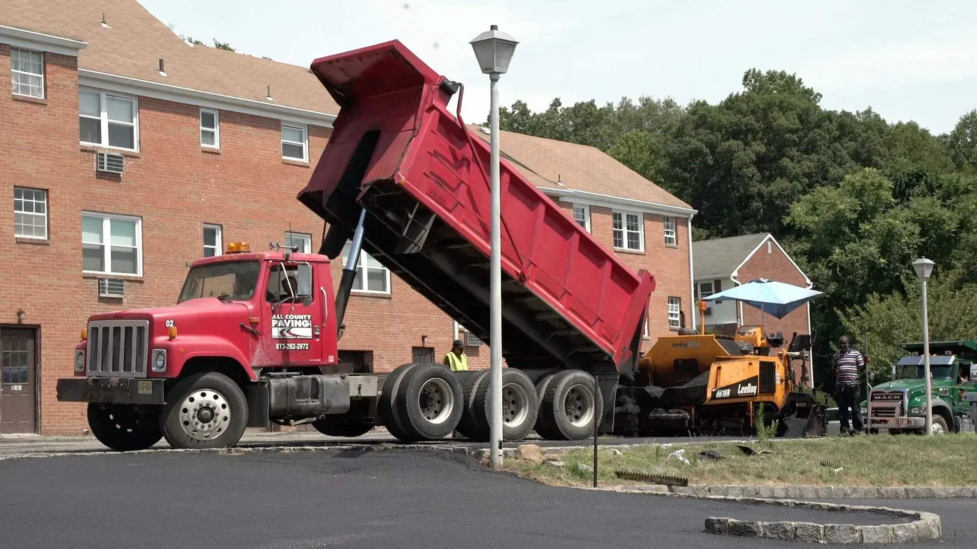 A red dump truck unloading asphalt into a paving machine in front of a brick apartment building.