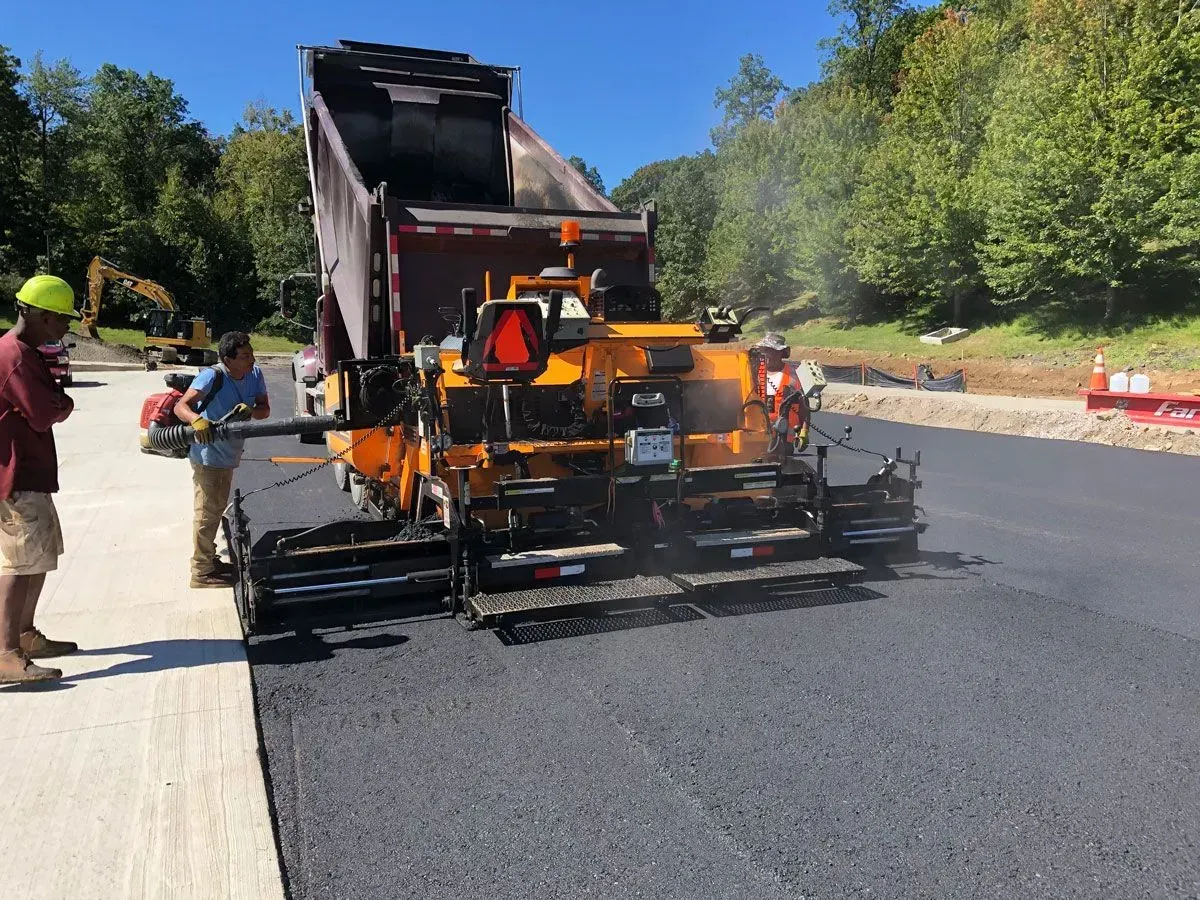 Asphalt paving in progress: A dump truck unloading asphalt into a paving machine, with workers nearby under a blue sky.