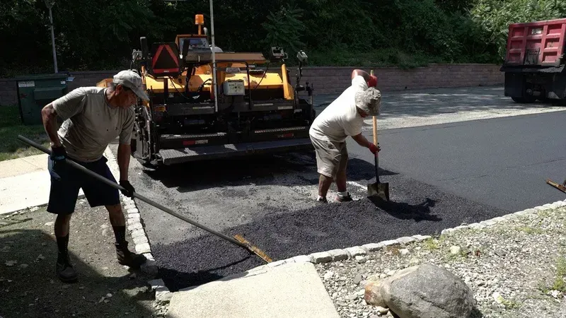 Two road workers paving asphalt on a street next to a curb, with a paving machine in the background.