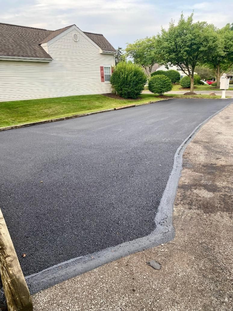 Newly paved asphalt driveway in front of a white house with a green lawn, trees, and a cloudy sky.