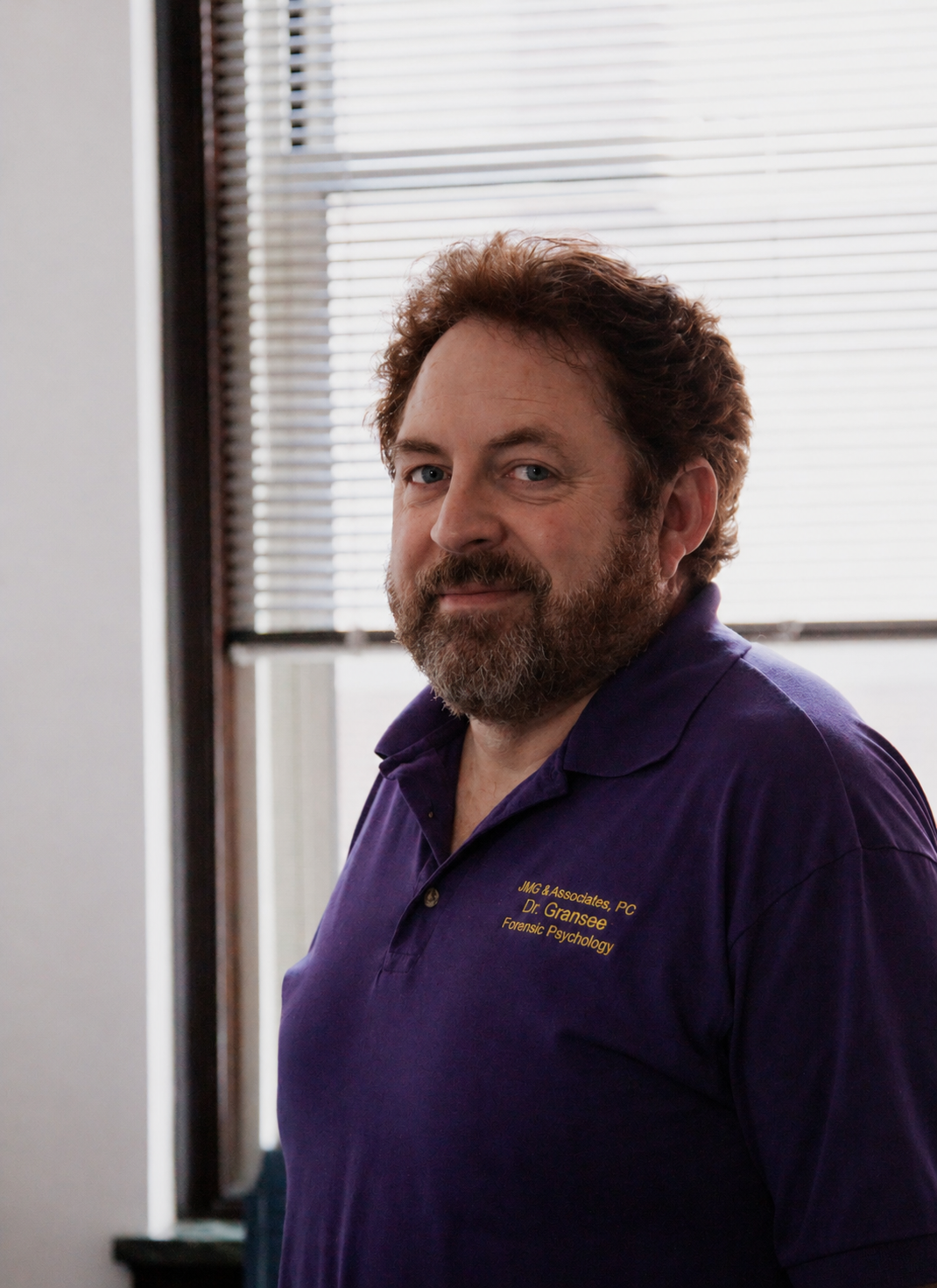 Man in a purple polo shirt standing by a window with blinds, looking at the camera.