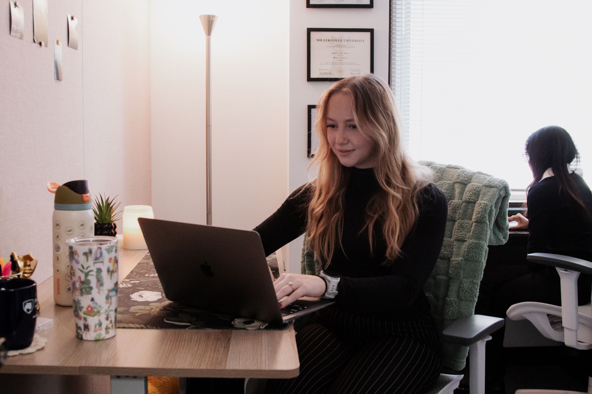 Woman working on a laptop at a desk in a bright office, with framed certificates on the wall.