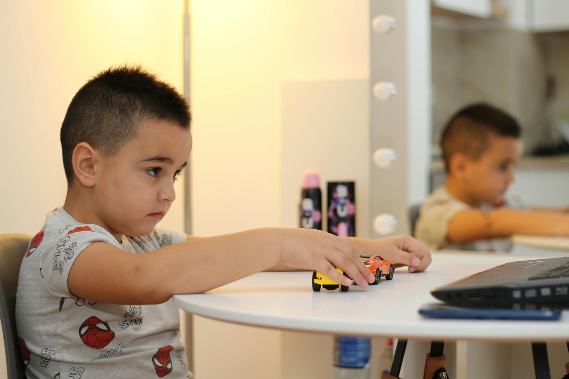 Child playing with toy cars at a white table, with a mirror reflecting the scene in the background