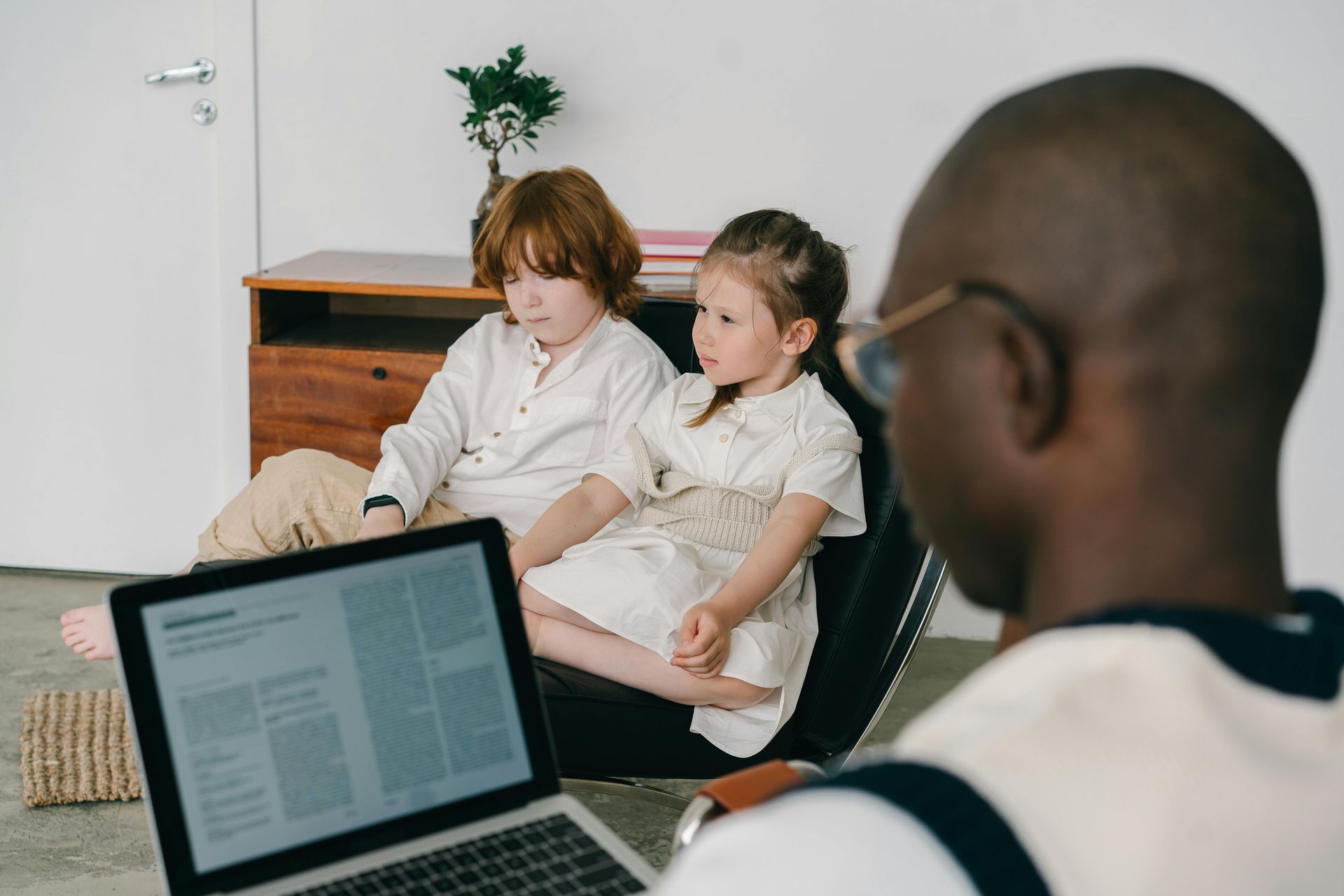 Three people in a living room, one holding a laptop while two sit on a couch listening.