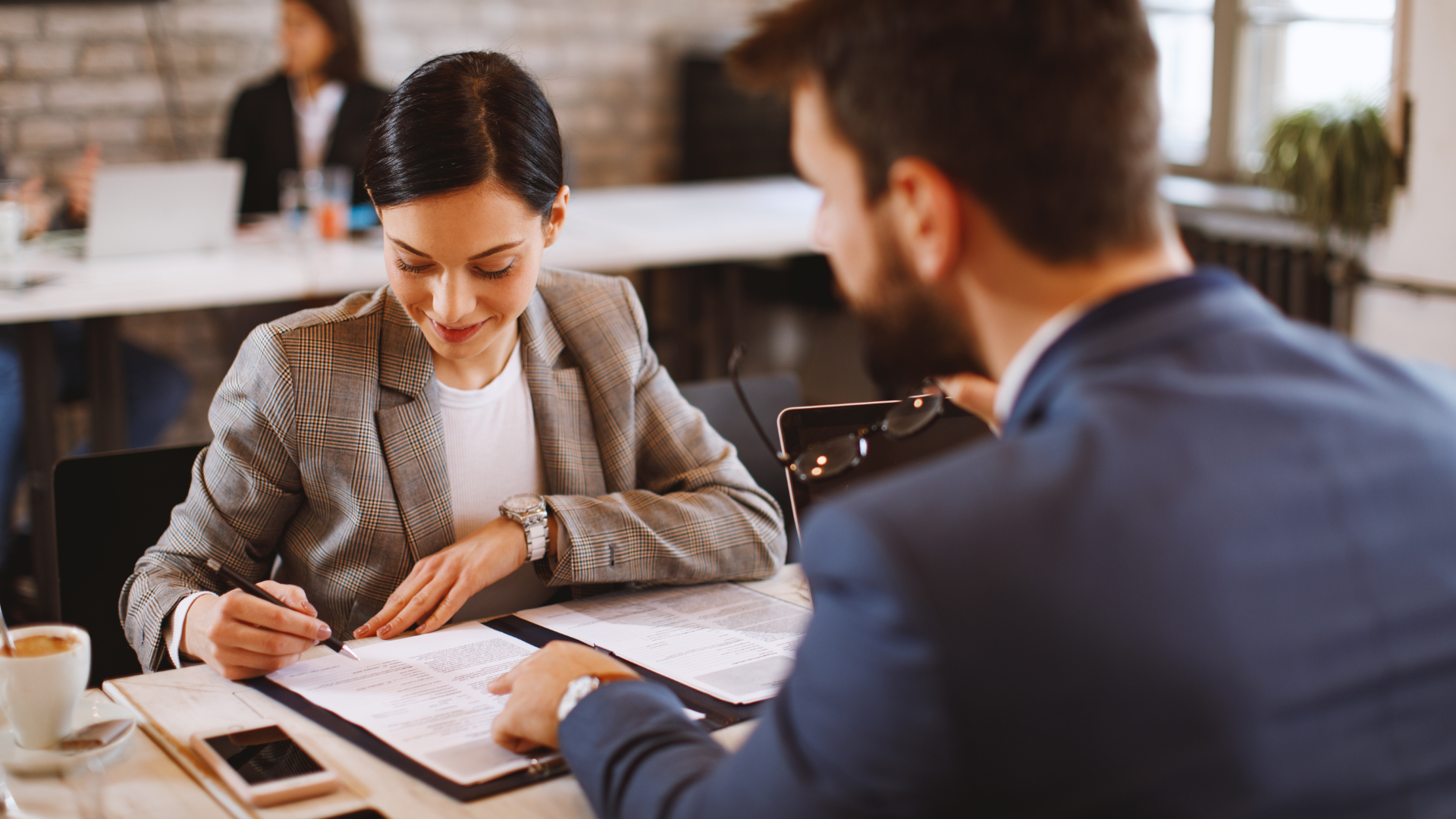 Two people reviewing documents at a table in a bright office, one writing and the other listening