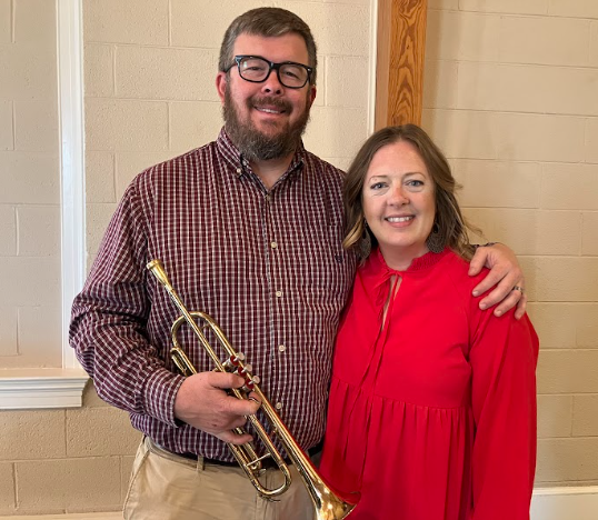 Man with trumpet and woman in red dress pose, smiling, indoors.