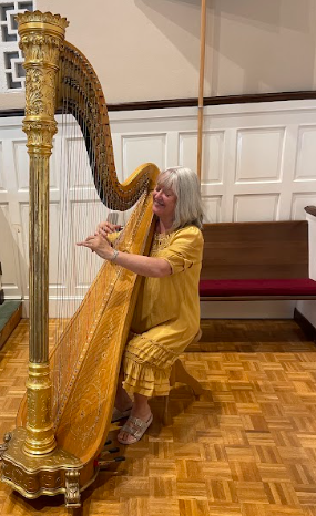 Woman playing a golden harp indoors; wearing yellow outfit, smiling.