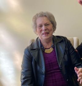 An elderly woman wearing a black leather jacket and a purple dress is sitting in a chair.