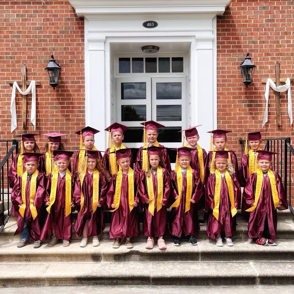 A group of children wearing graduation caps and gowns are standing in front of a brick building.